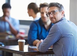 Two men having a conversation at a table whilst drinking coffee when the man nearest to the camera turns and looks at the camera.