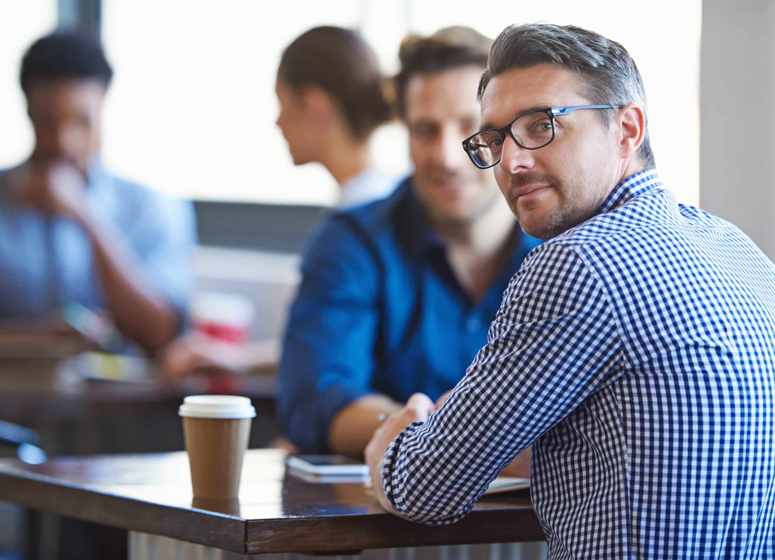 Two men having a conversation at a table whilst drinking coffee when the man nearest to the camera turns and looks at the camera.
